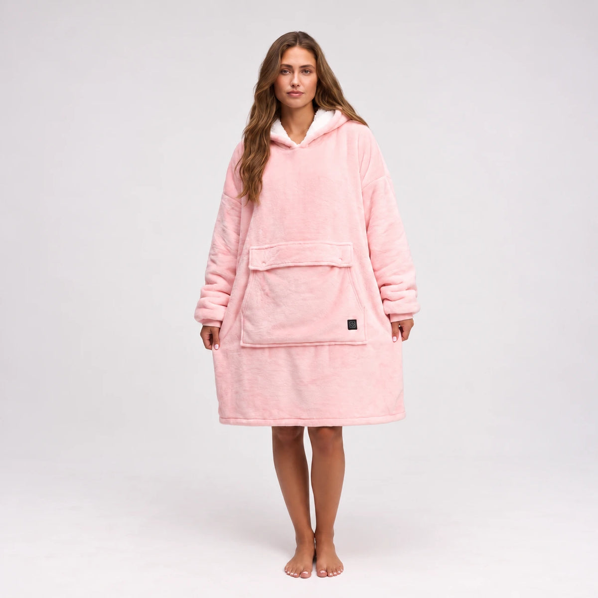 A woman stands barefoot against a light gray studio background wearing a plush Solrevé Solis™ hooded robe in a soft light pink color. The robe has a front kangaroo pocket and a small black square logo patch near the lower pocket.