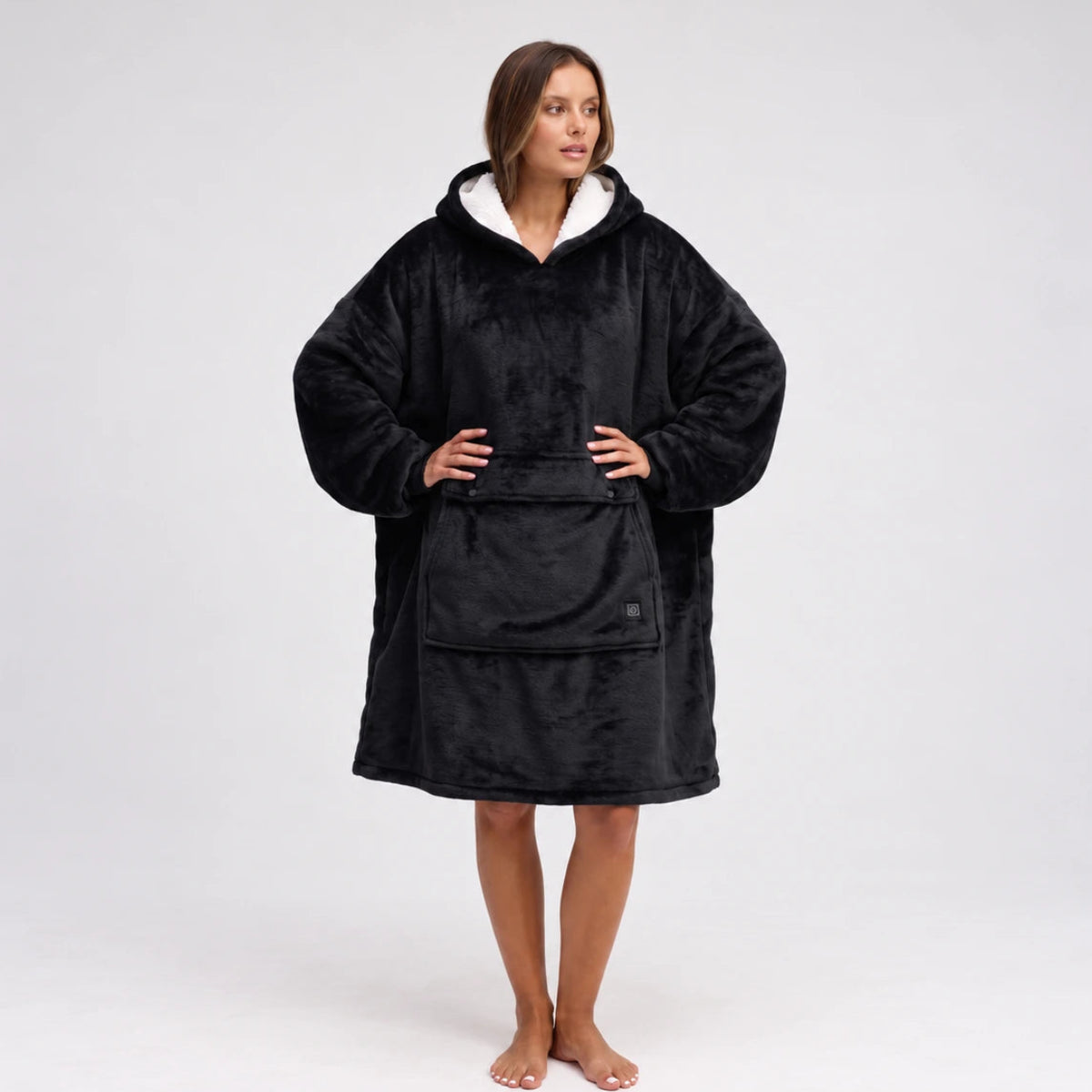 A woman modeling a Solrevé Solis™ black fleece hooded sweatshirt robe with a white-lined hood and a front kangaroo pocket, standing against a light gray studio background.