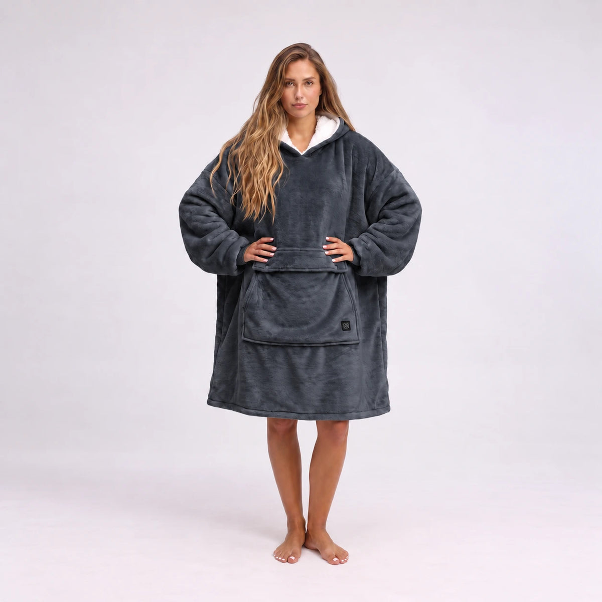 A woman stands barefoot against a light gray studio background wearing a Solrevé Solis™ dark charcoal gray hooded robe with a white inner hood lining and a large front patch pocket.