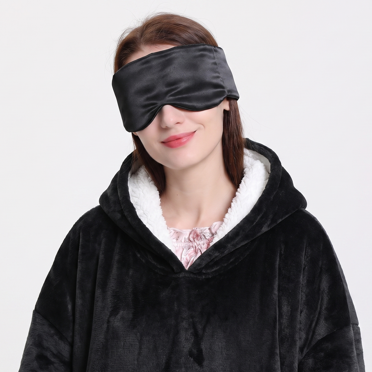A woman wearing a black velvet sleep mask and a black fleece hooded robe with a white sherpa lining, photographed against a plain light gray background.
