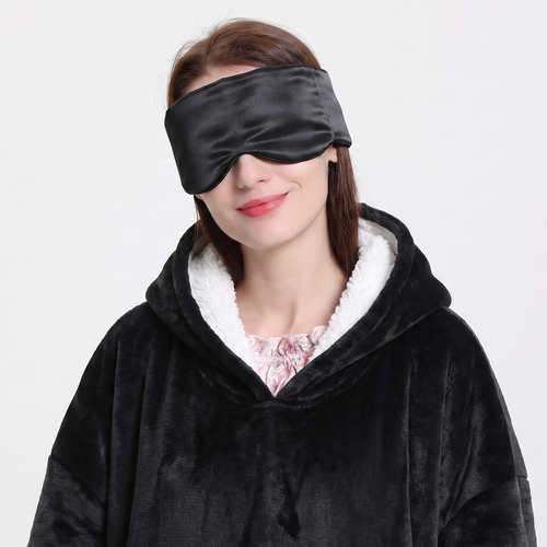 A woman wearing a black velvet sleep mask and a black fleece hooded robe with a white sherpa lining, photographed against a plain light gray background.